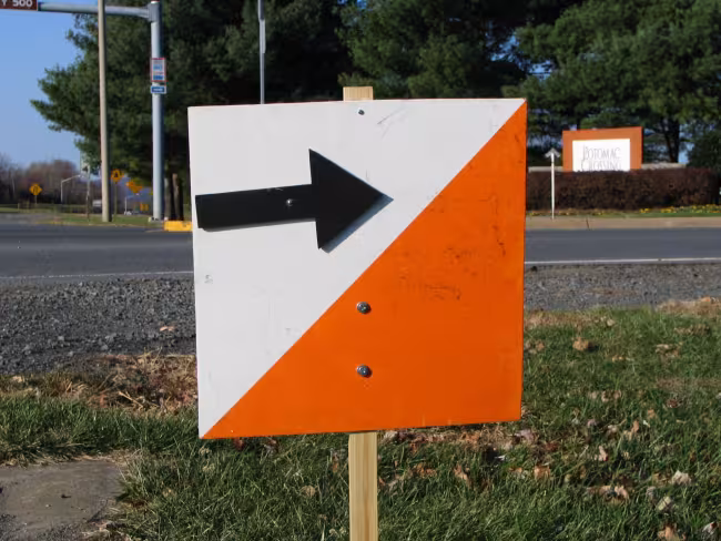 Orange-and-white orienteering sign with a black arrow, marking the turn to an event